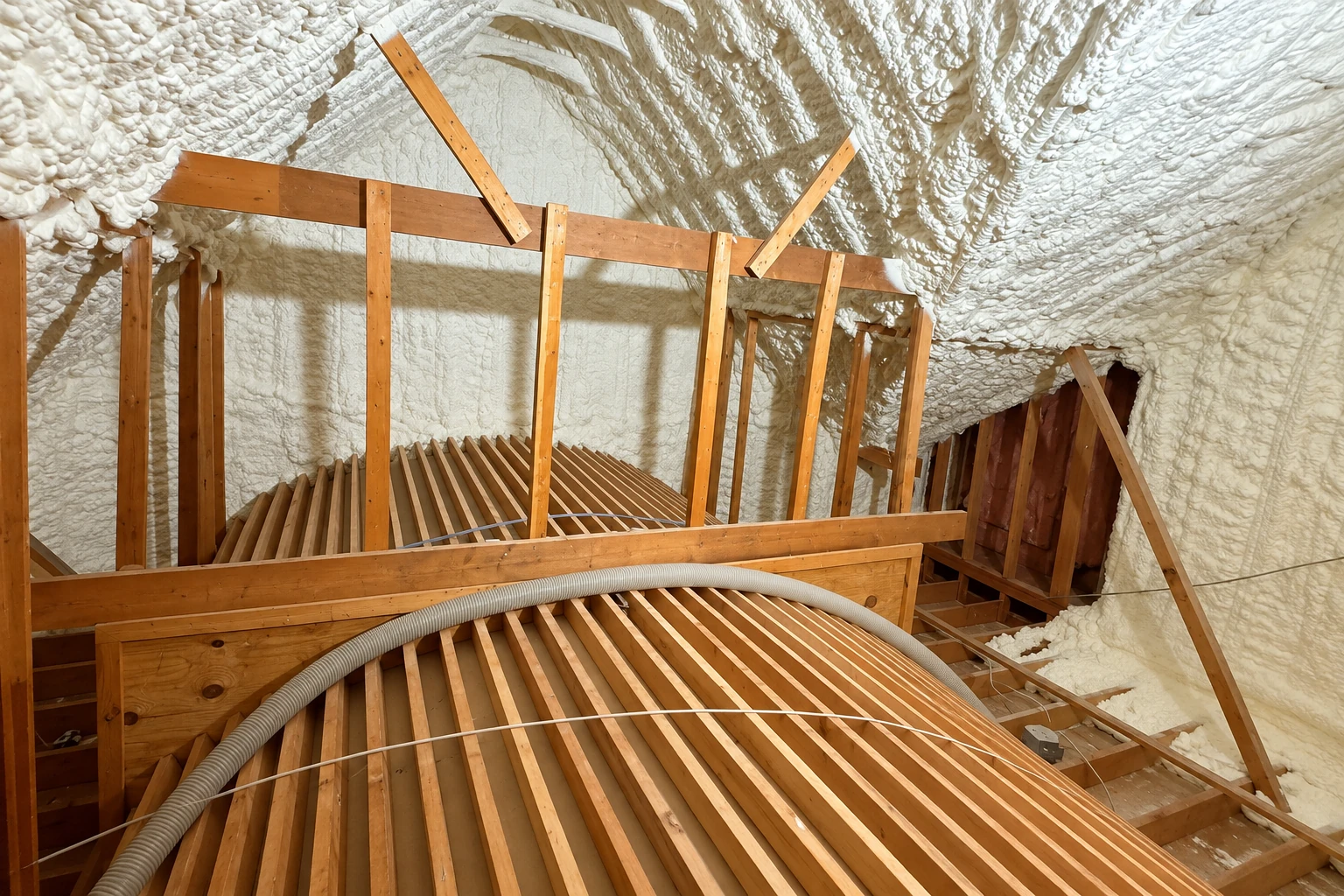 Attic rafters coated with white spray foam insulation, with exposed wooden framing and a flexible hose in the foreground.