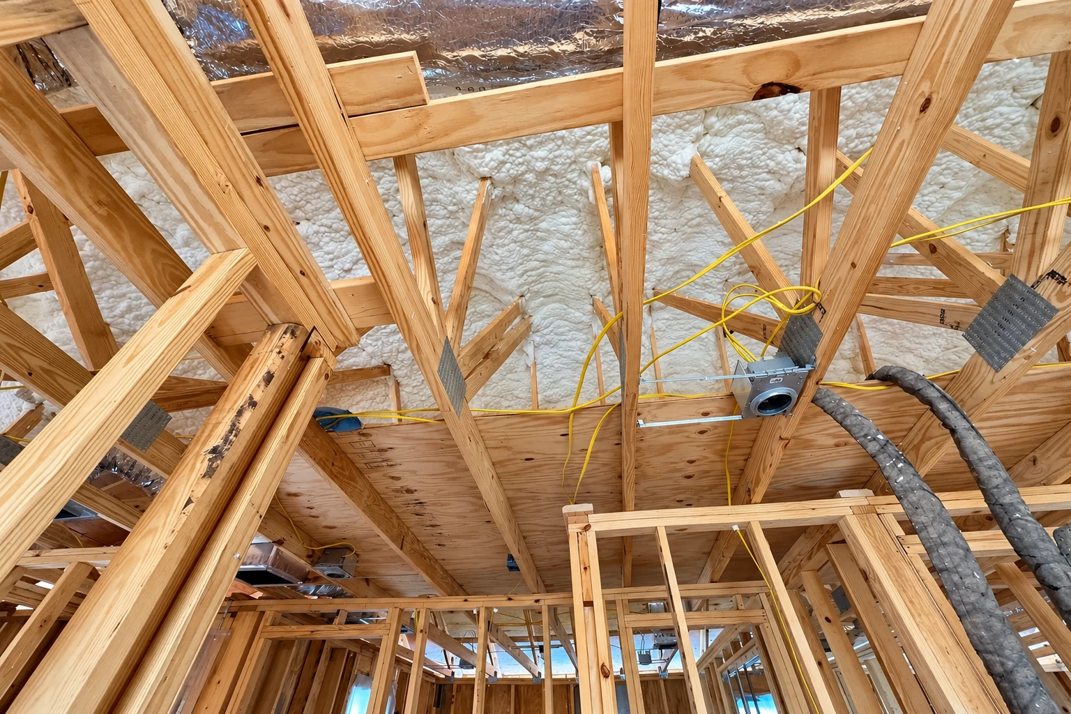 Exposed roof framing with white spray foam between rafters, yellow wiring, and a ventilation duct.