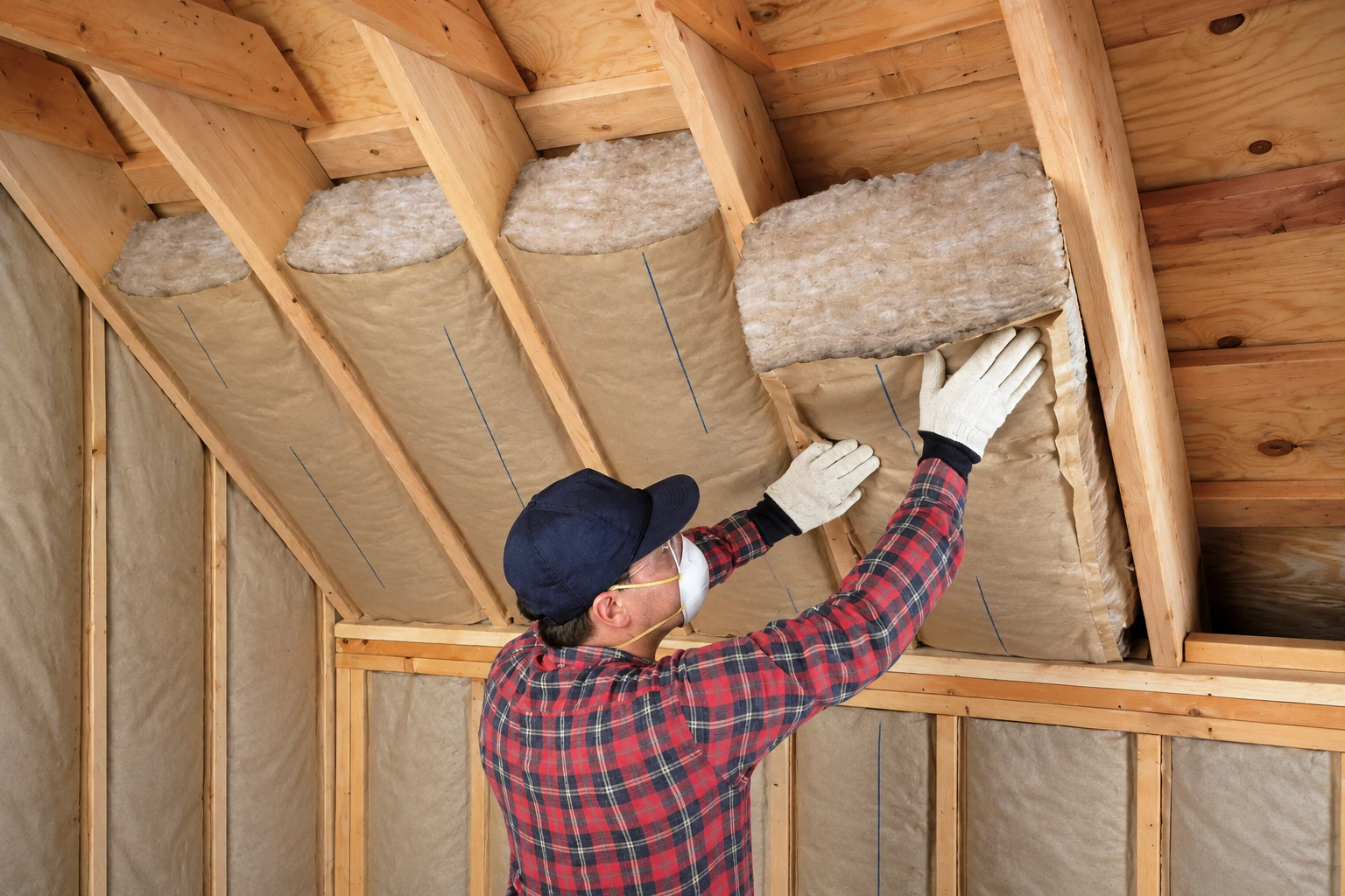 A masked worker fits fiberglass batts between the wooden rafters of an attic.