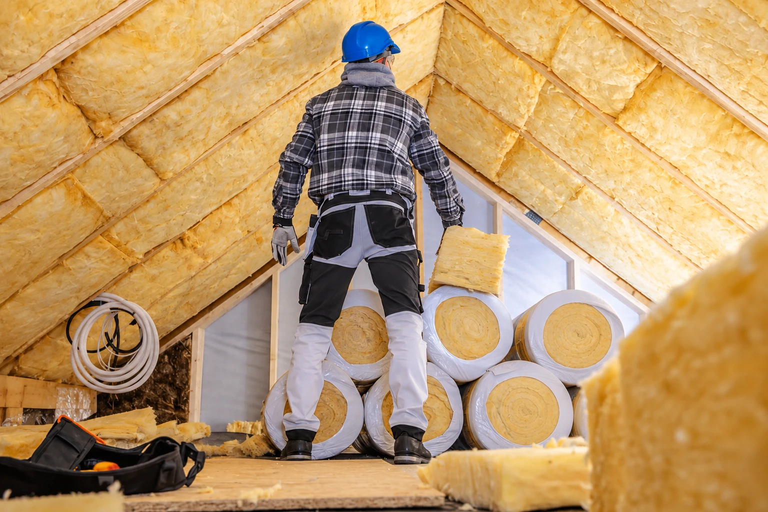 A worker in a blue hard hat stands among insulation rolls in an attic lined with yellow fiberglass batts.