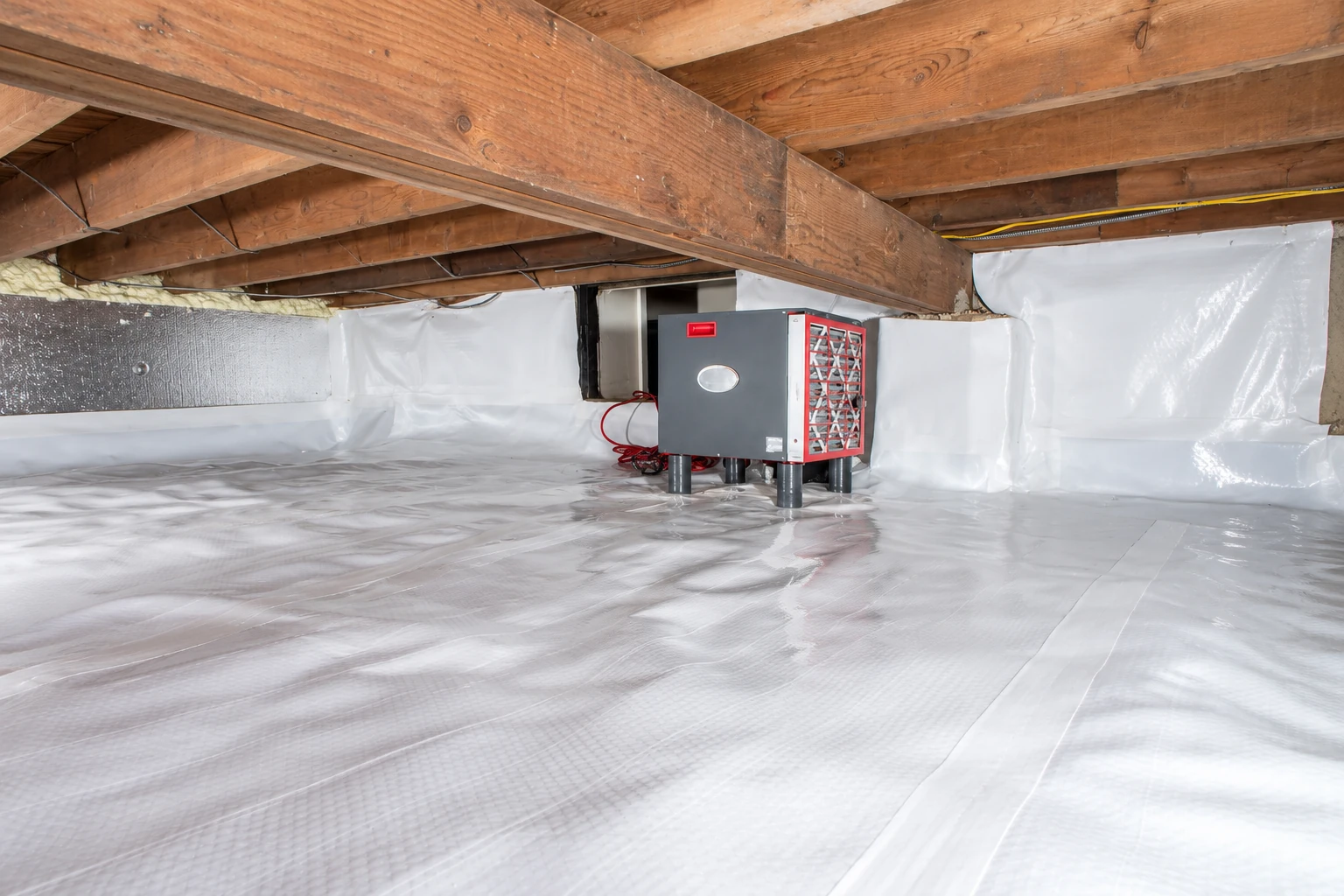 A crawl space floor and lower walls are covered with white vapor barrier beneath wooden floor joists, with a utility unit at the back.
